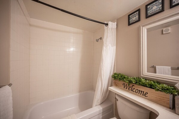 Un baño con una bañera blanca, una cortina de ducha, un letrero de 'Bienvenido', un espejo y una planta pequeña en el mostrador.
