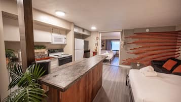 A modern hotel room with a kitchenette, a bed with a white comforter, and a view into the adjacent room.