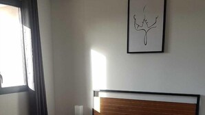 Ein Schlafzimmer mit einem Bett, einem Nachttisch, einer Lampe, einem gerahmten Kunstwerk und einem Fenster mit Vorhängen.