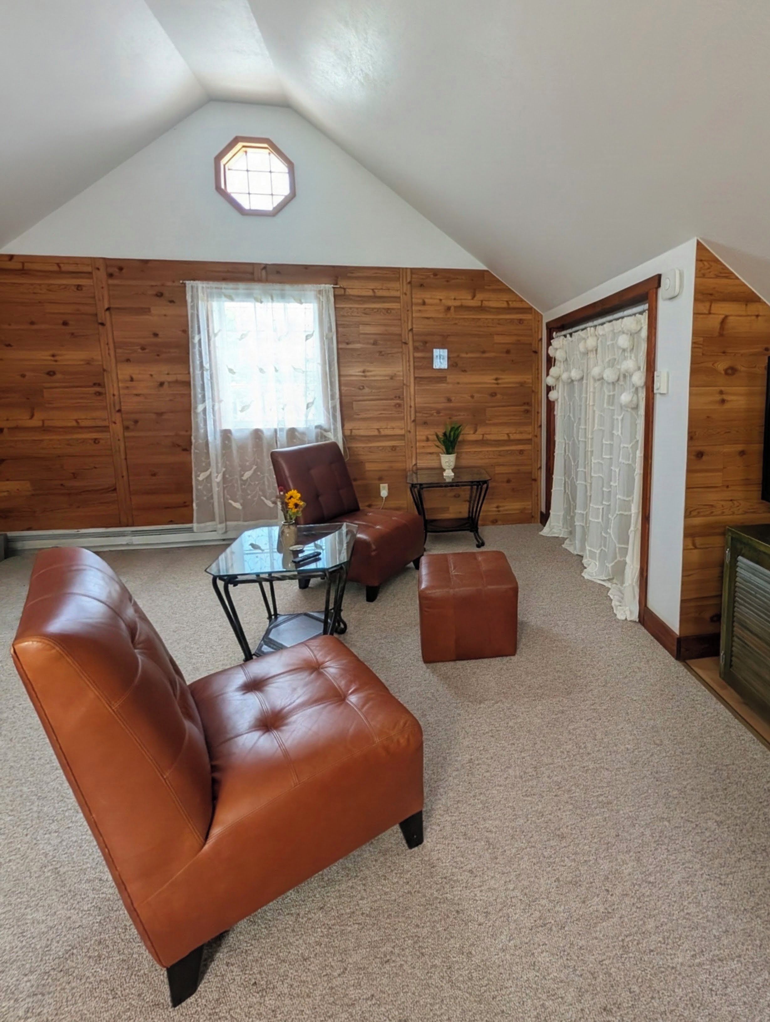 A loft-style room with a sloped ceiling, a large window, a brown leather sofa, a glass coffee table, and a small side table.