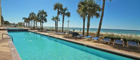 A swimming pool with clear blue water, surrounded by palm trees and lounge chairs, with a beach and ocean in the background.
