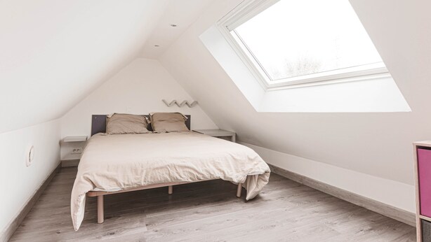 A bedroom with a sloped ceiling, a skylight, a bed with a beige comforter, and a nightstand with a lamp.