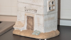 Une petite décoration en bois, de couleur blanche, en forme de maison, sur laquelle est inscrit le mot « Été ».