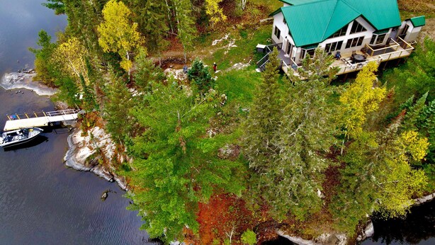 An aerial view of a lakeside property with a house featuring a green roof, a dock with a boat, and surrounding dense forest.