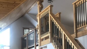 A wooden staircase with a balustrade, a wooden rocking chair, a small table, and a lamp in a room with a high ceiling and windows.
