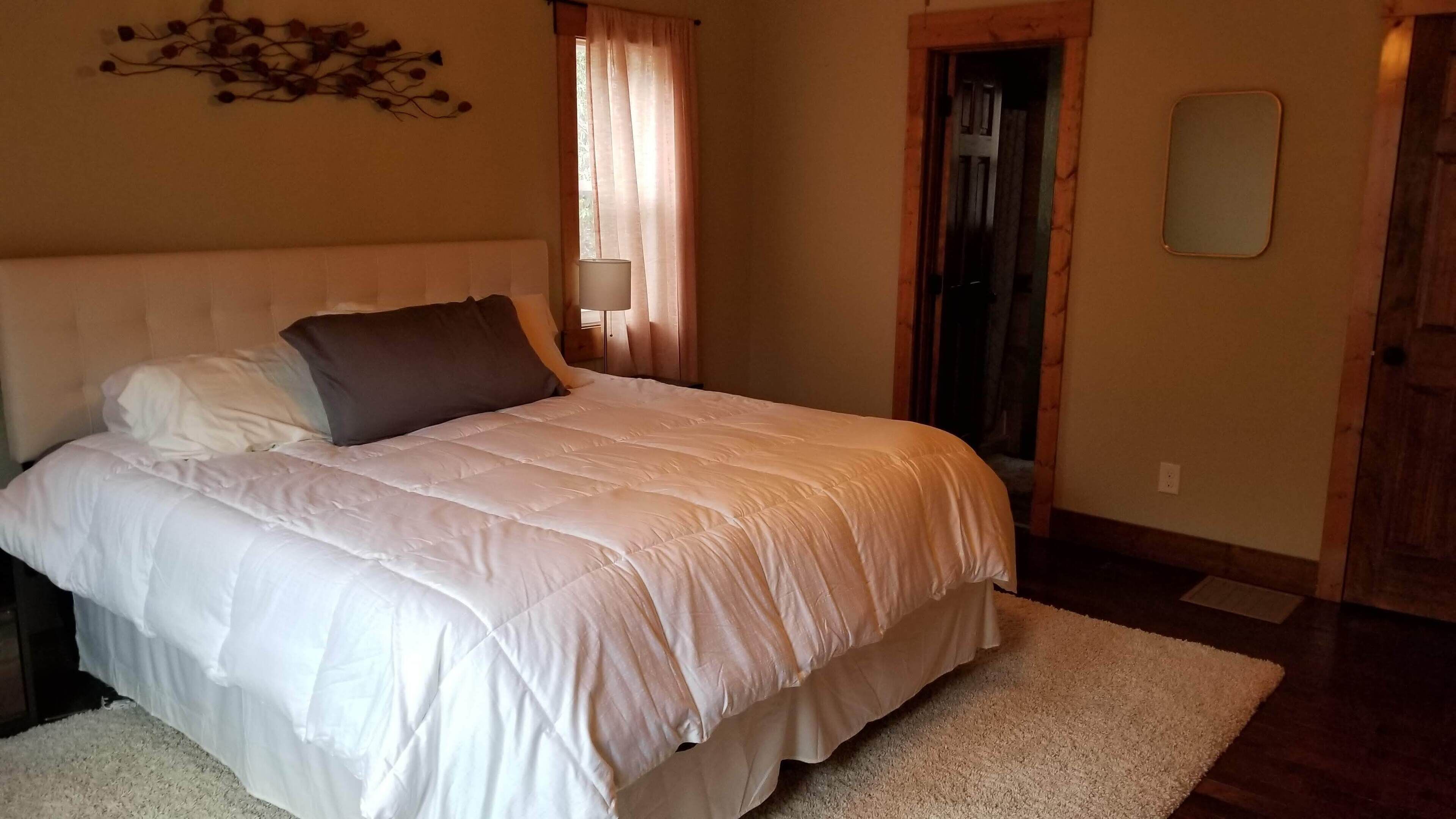 A bedroom with a large bed, a nightstand, a wall-mounted mirror, and a door leading to another room.