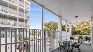 A covered balcony with a view of a parking lot and trees, furnished with a table and chairs.