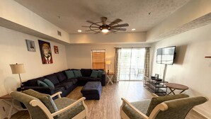 A living room with a sectional sofa, two armchairs, a coffee table, a ceiling fan, a TV, and a sliding glass door leading to a balcony.