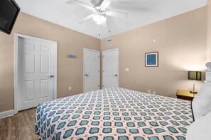 A bedroom with a patterned bedspread, two white doors, a ceiling fan, a nightstand with a lamp, and a framed picture on the wall. - Hum-Dinger, pool, lazy river & 2 hot tubs, Family friendly, 200yards to beach! (Myrtle Beach)