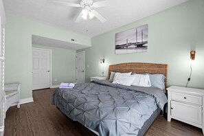 A bedroom with a large bed, a ceiling fan, a nightstand, a wicker chair, and a wall-mounted lamp. - Cloud 9-er (Myrtle Beach)