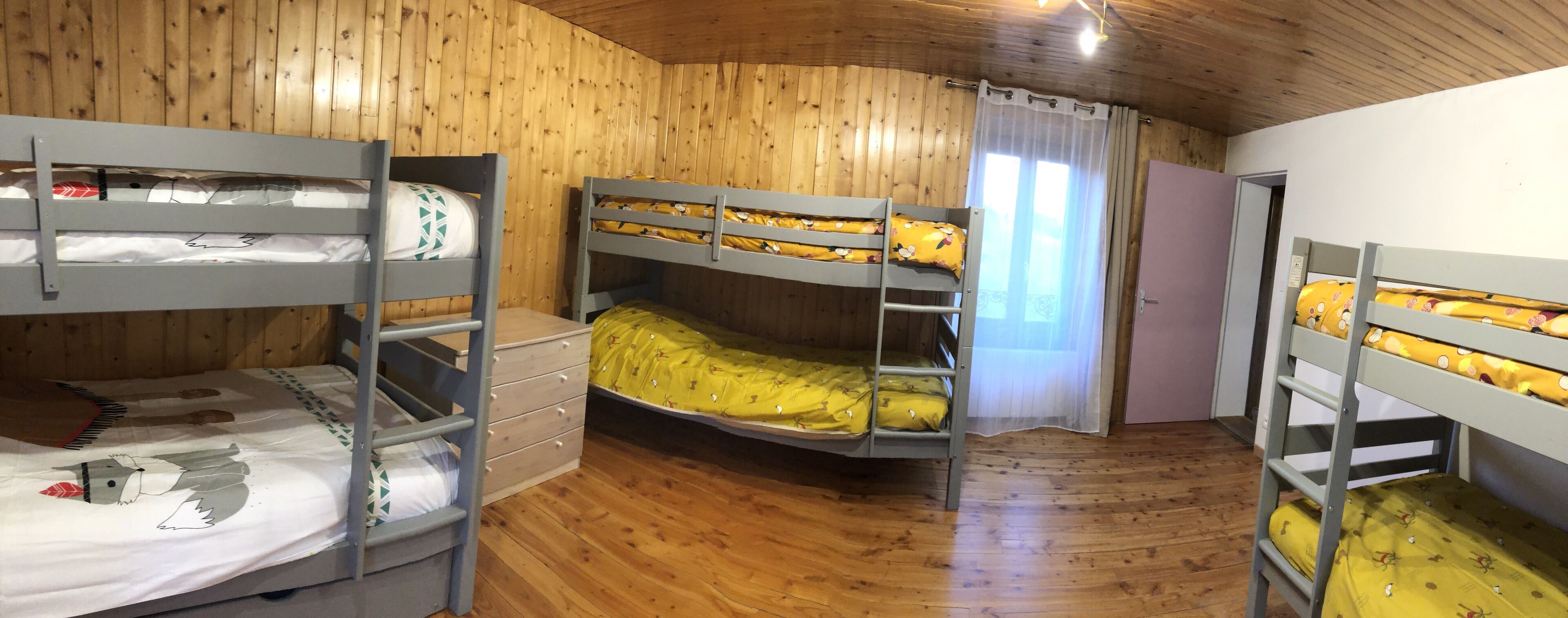 A room with wooden walls and floors, bunk beds with yellow bedding, a white door, and a window with curtains.