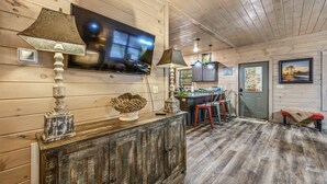 Un interior rústic amb un paviment de fusta, un televisor muntat a la paret, un armari de fusta i una zona de menjador amb caires i una taula.
