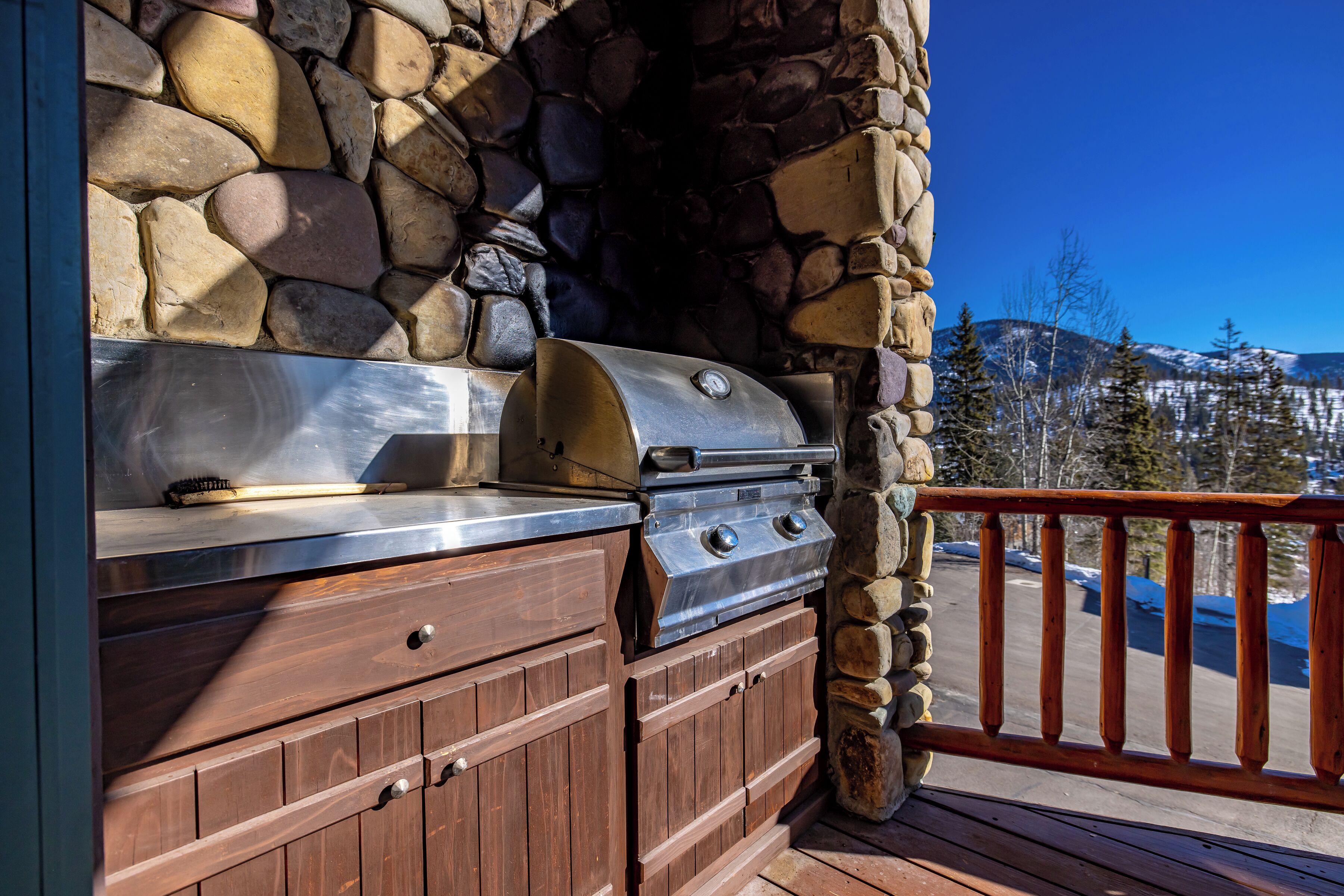 An outdoor stone fireplace with a metal grill, wooden cabinets, and a wooden deck railing.
