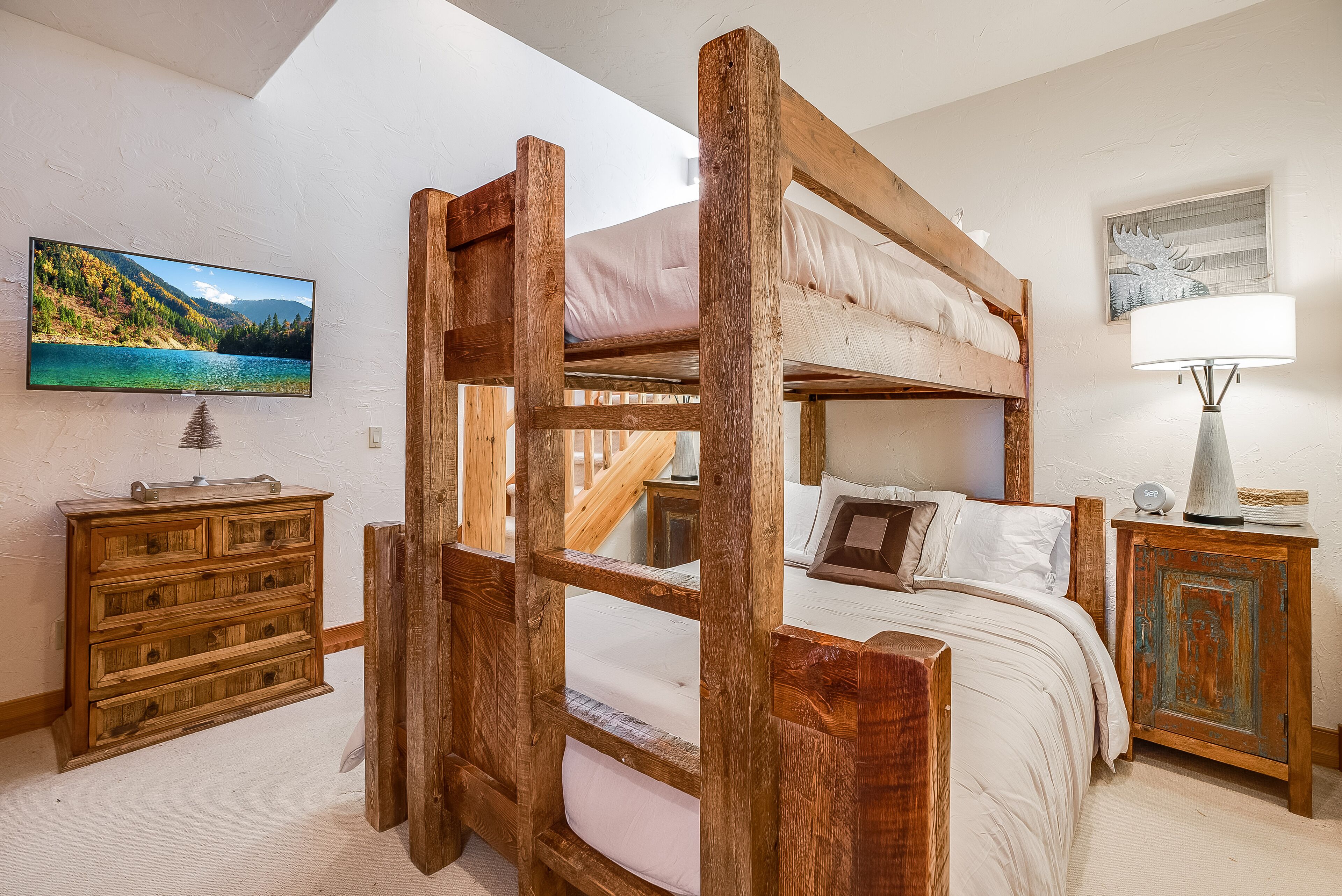 A bunk bed with a wooden frame, a television mounted on the wall, a wooden dresser, and a lamp on a side table.