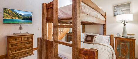 A bunk bed with a wooden frame, a television mounted on the wall, a wooden dresser, and a lamp on a side table.