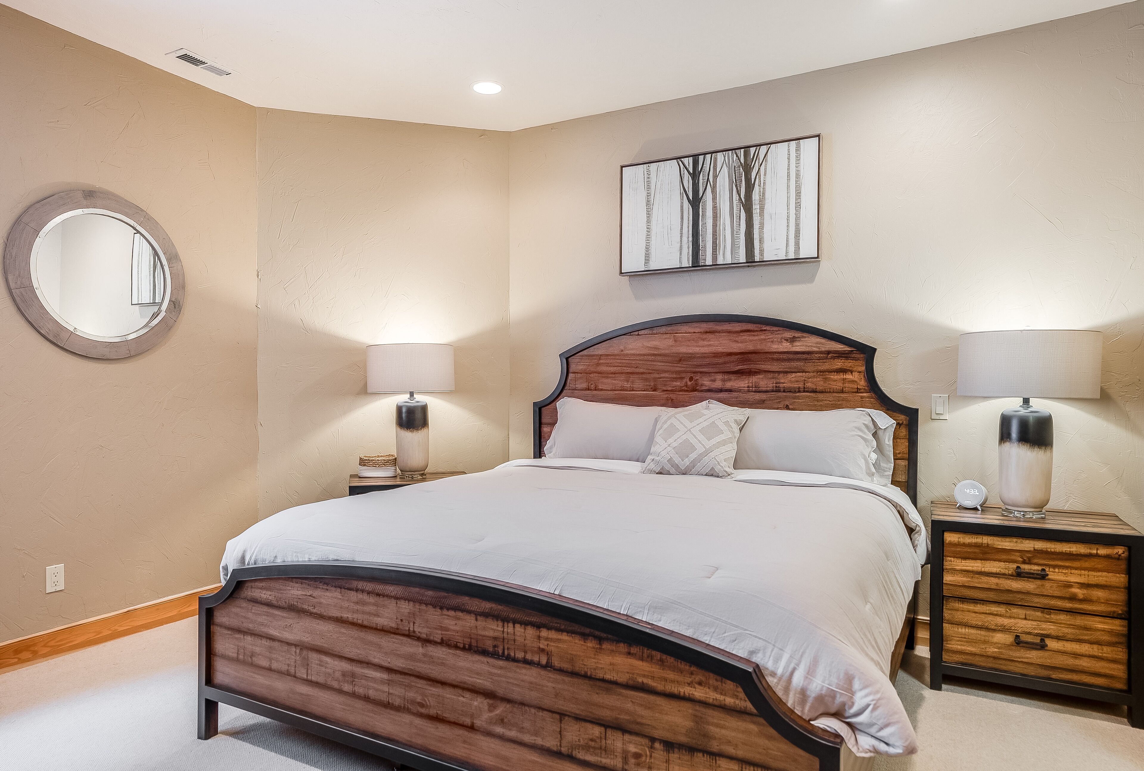 Un dormitorio con estructura de cama de madera, dos mesitas de noche con lámparas, un espejo redondo y una obra de arte colgada en la pared.