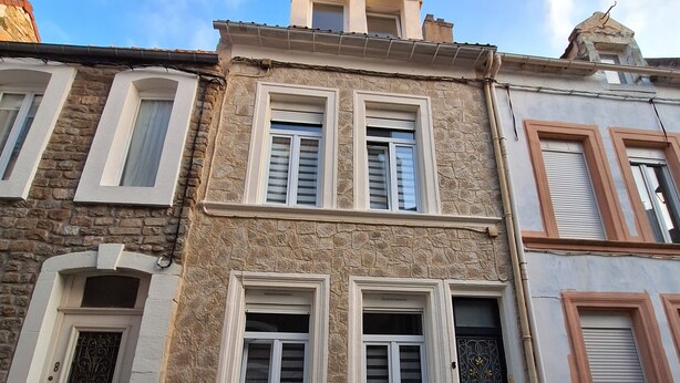 Un bâtiment à deux étages avec une façade en pierre, de nombreuses fenêtres avec volets et une rue pavée.