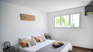 A bedroom with a bed, bedside tables, a TV, and a window with a view of palm trees. - La Maline**** Piscine chauffée et salle de jeux (Sainte-Soulle)