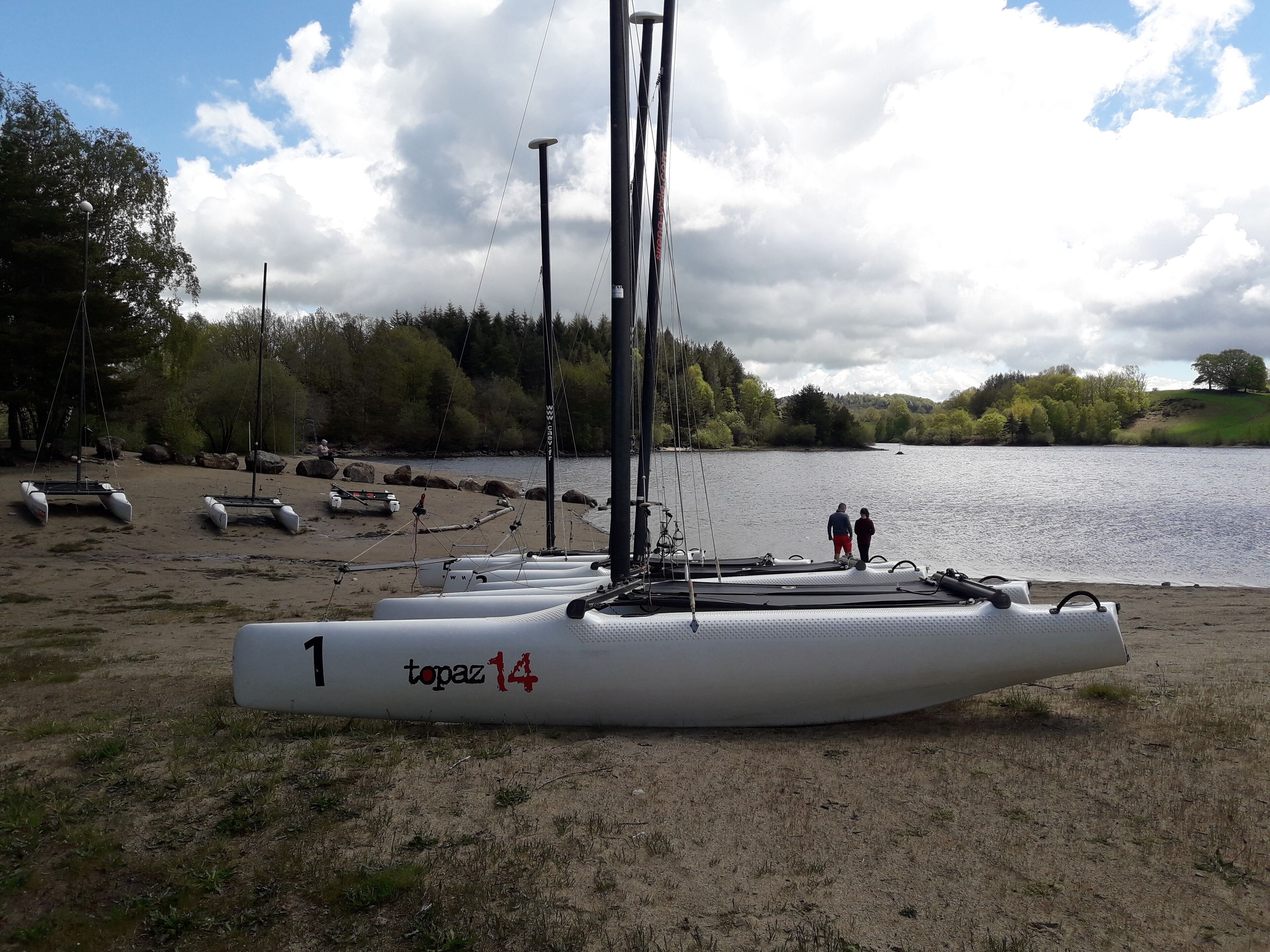 Ein Segelboot namens 'topaz 14' an einem Sandstrand mit einem stillen See und Bäumen im Hintergrund.