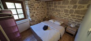 A bedroom with a stone wall, wooden floor, a bed with white bedding, a nightstand, and a window with curtains.