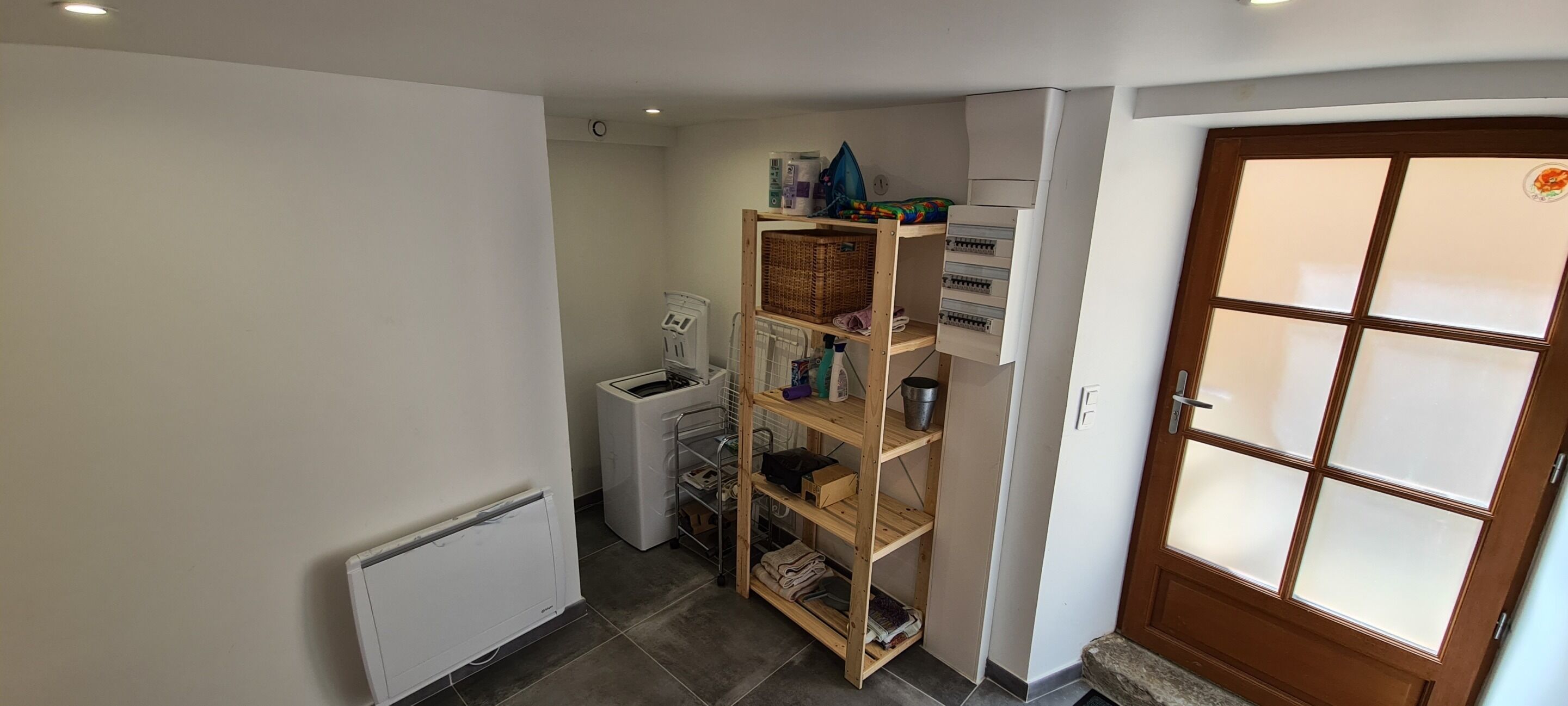 Un couloir avec une étagère en bois, un radiateur blanc, un appareil électroménager blanc et une porte dotée de panneaux en verre d époli.