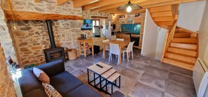 A cozy living room with a stone wall, a sofa, a coffee table, and a TV. There's a kitchen area with wooden cabinets and a dining table.