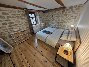 Een slaapkamer met een stenen muur, houten vloer, een bed met kussens en dekbed, een nachtkastje met lamp, en een raam met gordijnen.