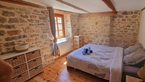A bedroom with stone walls, a wooden floor, a bed with a leaf-patterned bedspread, and a window with curtains.