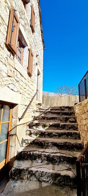 Un escalier en pierre mène à un bâtiment doté de volets en bois et d’une rambarde en métal.