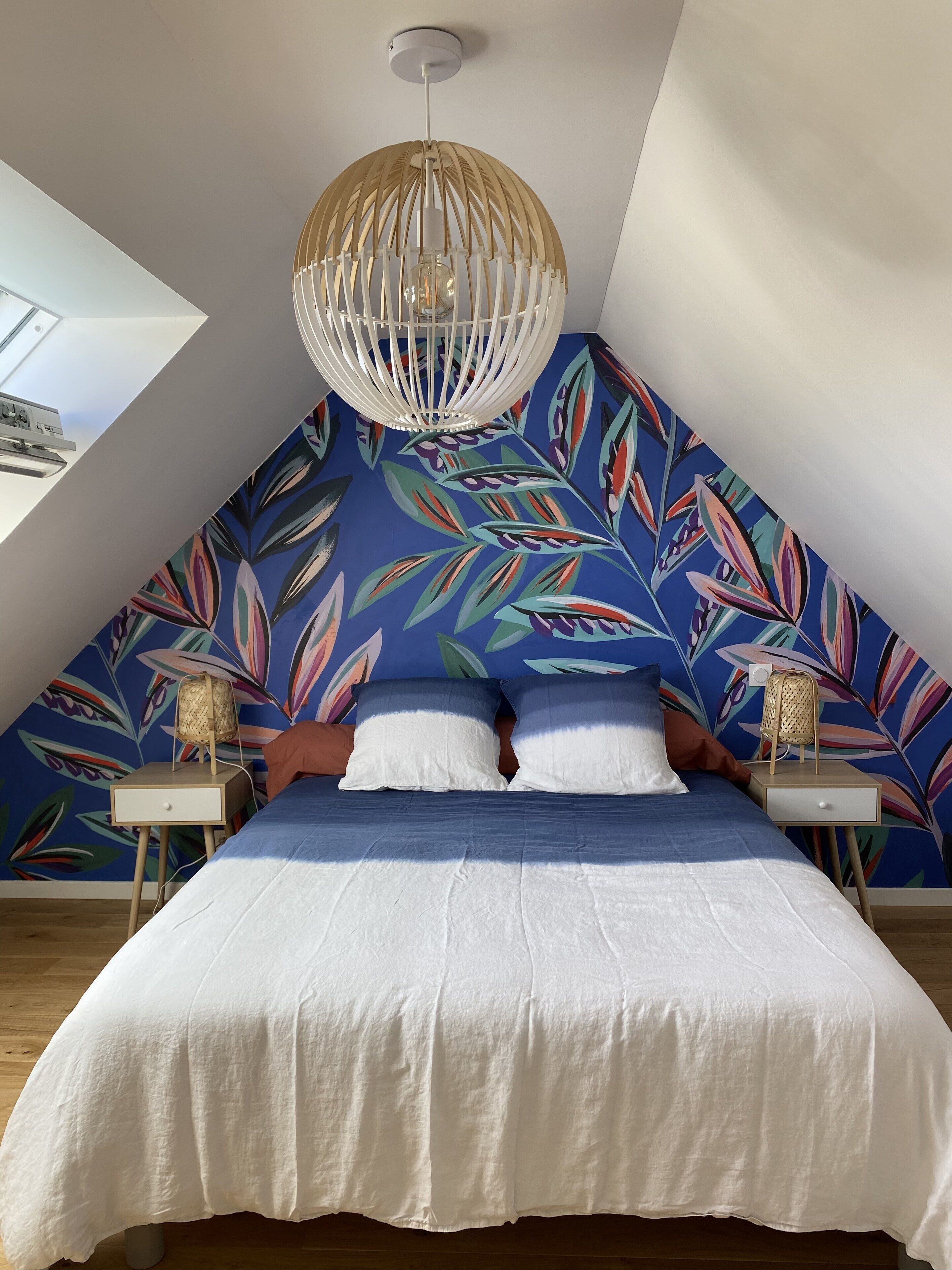 Een slaapkamer met een schuine plafond, een groot bed met wit beddengoed, twee nachtkastjes met lampen, en een muur met een levendig bladpatroon.