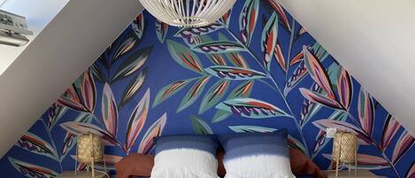 Een slaapkamer met een schuine plafond, een groot bed met wit beddengoed, twee nachtkastjes met lampen, en een muur met een levendig bladpatroon.