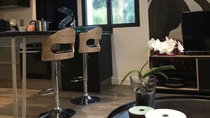 A modern kitchen with two bar stools, a dining table with decorative items, and a large window with a view of greenery. - L' Annex ' : charming quiet house with garden close to the beach (Névez)