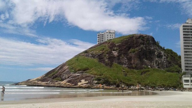 A beach with a rocky hill, a building on top, and a tall building to the right. - Penthouse with its own barbecue near Enseada Beach - Guarujá (Guarujá)