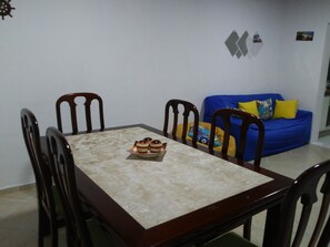 A dining area with a rectangular table and four chairs, a blue sofa with yellow cushions, and a wall with geometric decorations. - Penthouse with its own barbecue near Enseada Beach - Guarujá (Guarujá)