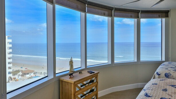 Un dormitorio con un amplio ventanal con vistas a la playa, un aparador de madera y una cama con colcha de motivos náuticos.