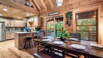 A wooden cabin interior with a dining area, kitchen, and a view of the outdoors.
