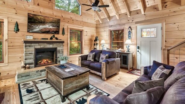 A log cabin interior with a fireplace, leather sofas, a ceiling fan, and a large window.