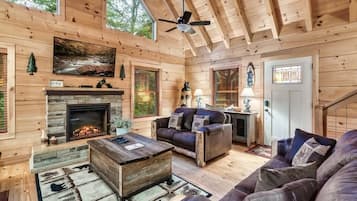 A log cabin interior with a fireplace, leather sofas, a ceiling fan, and a large window.