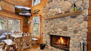 Interior rústico de una cabaña con chimenea de piedra, muebles de madera y un televisor montado en la pared.