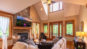 A spacious living room with a stone fireplace, comfortable seating, a flat-screen TV, and large windows offering a view of the outdoors.