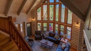 Ein Blockhaus-Interieur mit Schlafzimmer im oberen Bereich, einem Wohnzimmer mit Sofa und Couchtisch und großen Fenstern mit Blick ins Freie.