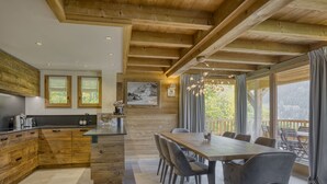 Une cuisine moderne avec des meubles en bois et un îlot central, un coin repas avec une grande table et des chaises, et de grandes fenêtres avec des rideaux.