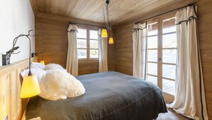 Une chambre dans une cabane en bois, avec un lit, deux fenêtres avec des rideaux et des lampes suspendues.