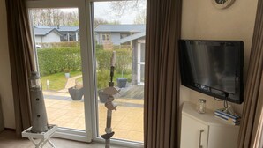 Una habitación con una televisión sobre un mueble blanco, una ventana grande con cortinas, una mesita con una vela y pañuelos de papel, y una escultura decorativa de pez en el suelo.