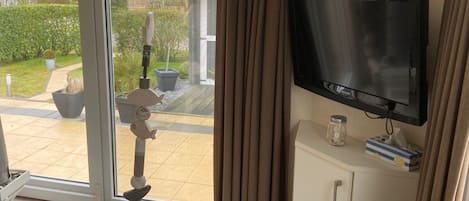 Una habitación con una televisión sobre un mueble blanco, una ventana grande con cortinas, una mesita con una vela y pañuelos de papel, y una escultura decorativa de pez en el suelo.