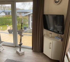 Een kamer met een televisie op een witte kast, een groot raam met gordijnen, een tafeltje met een kaars en zakdoekjes, en een decoratief vissenbeeld op de vloer.