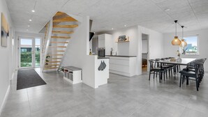 A modern kitchen with a central island, a dining area with a long table and chairs, and a staircase leading to an upper level. - Lovely home in Thisted (Thisted)
