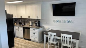 Una cocina moderna con armarios blancos, un refrigerador negro, lavavajillas y cafetera. Hay una mesa de comedor con dos sillas blancas y un televisor de pantalla plana montado en la pared.