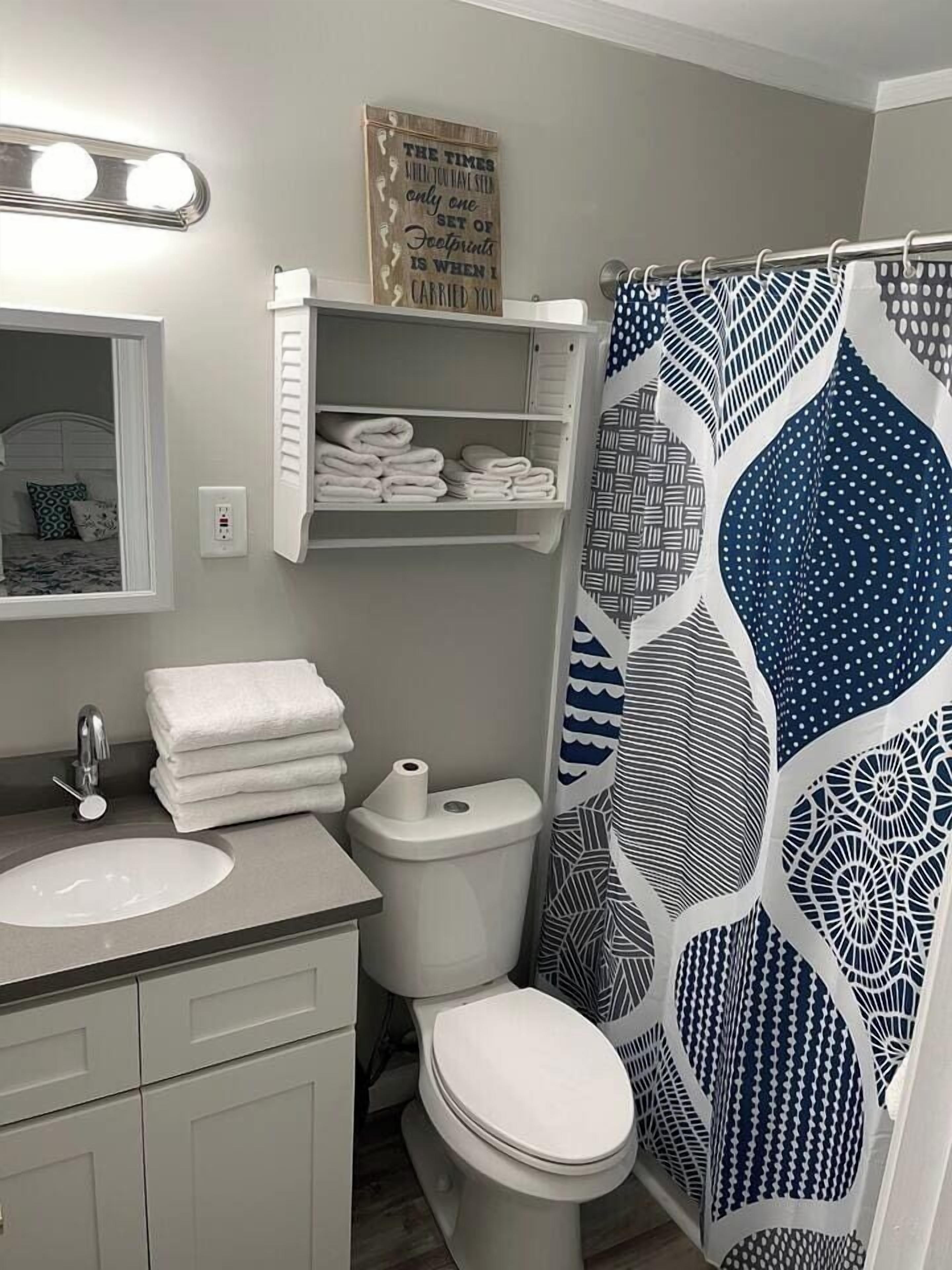 Un baño con un lavamanos blanco, un inodoro, un perchero con toallas dobladas y una cortina de ducha con un diseño geométrico.