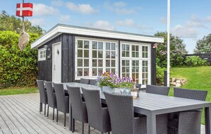 Um deck de madeira com uma mesa e cadeiras, um pequeno edifício com janelas amplas e uma bandeira.